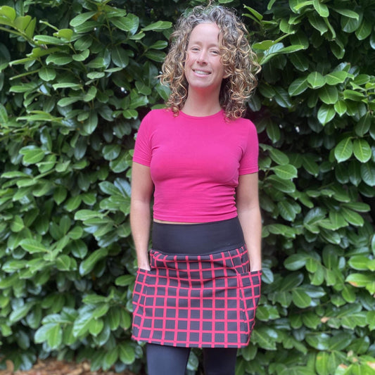 Woman wearing a pink top and red plaid skirt standing in front of a green hedge.