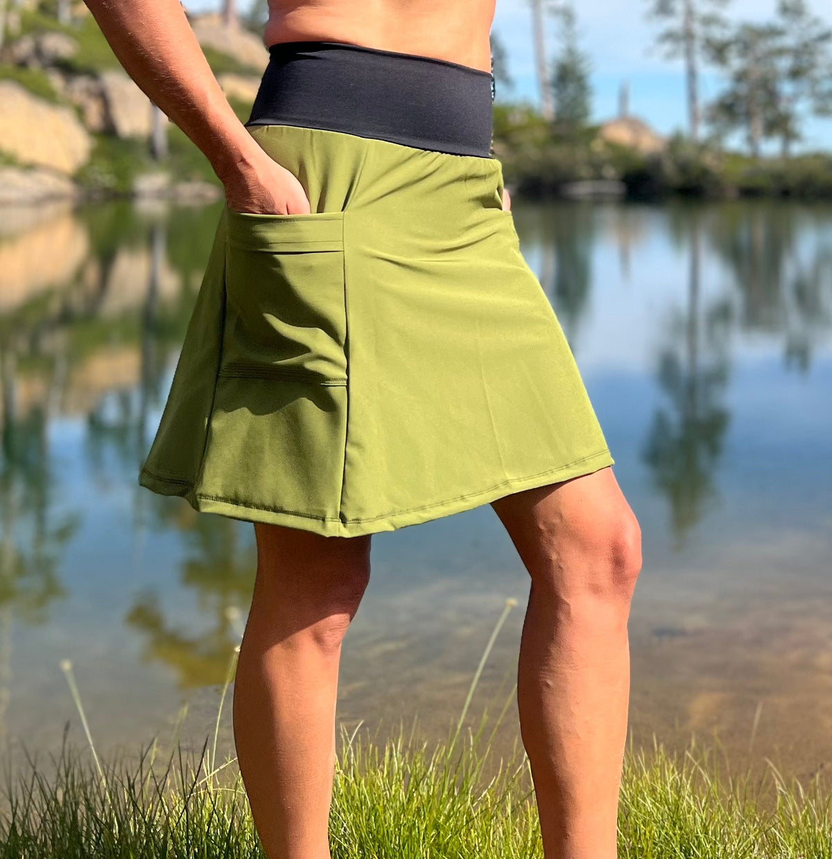 Person wearing a green Adventure Skirt with a wide black waistband and a hand in the hip pocket beside a lake.
