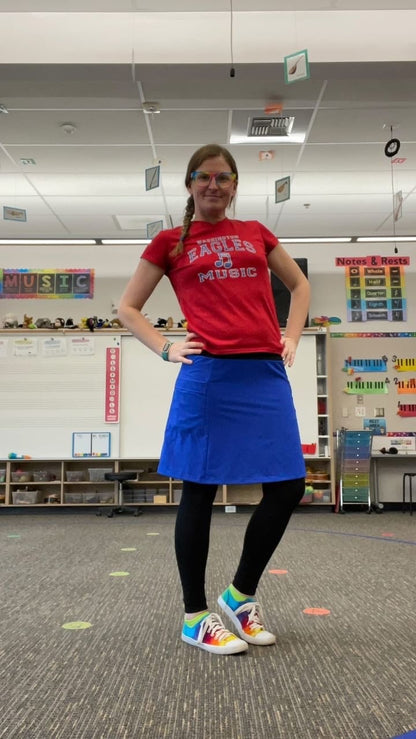 Person wearing a red shirt with 'Music' text, blue skirt, and colorful shoes in a classroom setting.