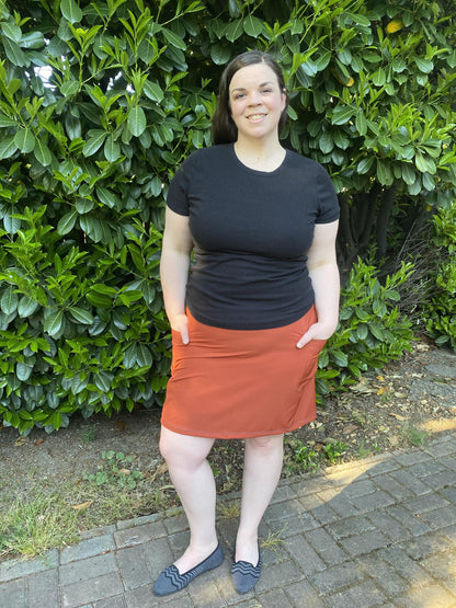 Woman wearing a black top and rust skirt standing in front of green bushes.