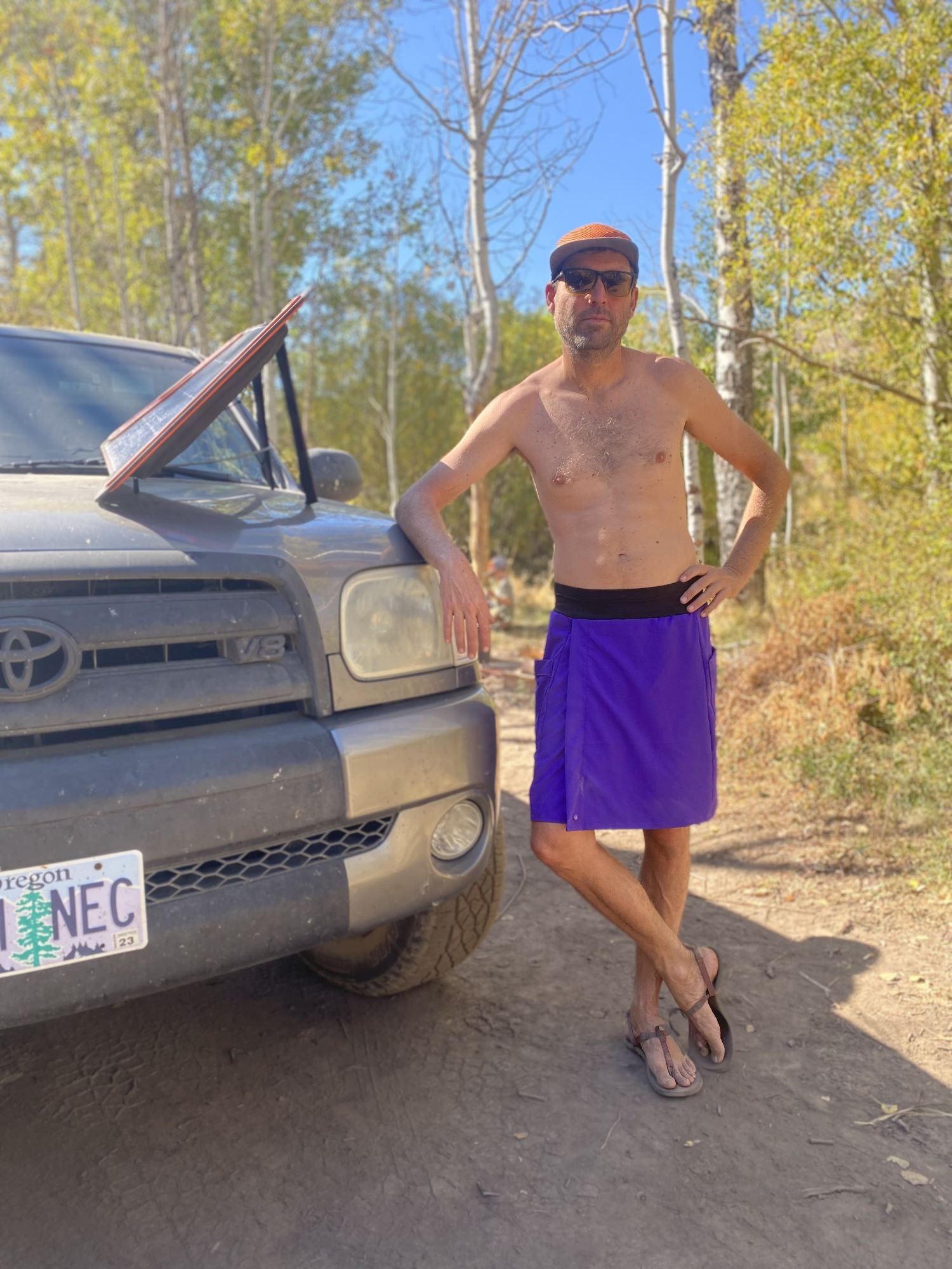 Man in a purple skirt standing next to a Toyota truck in a forested area