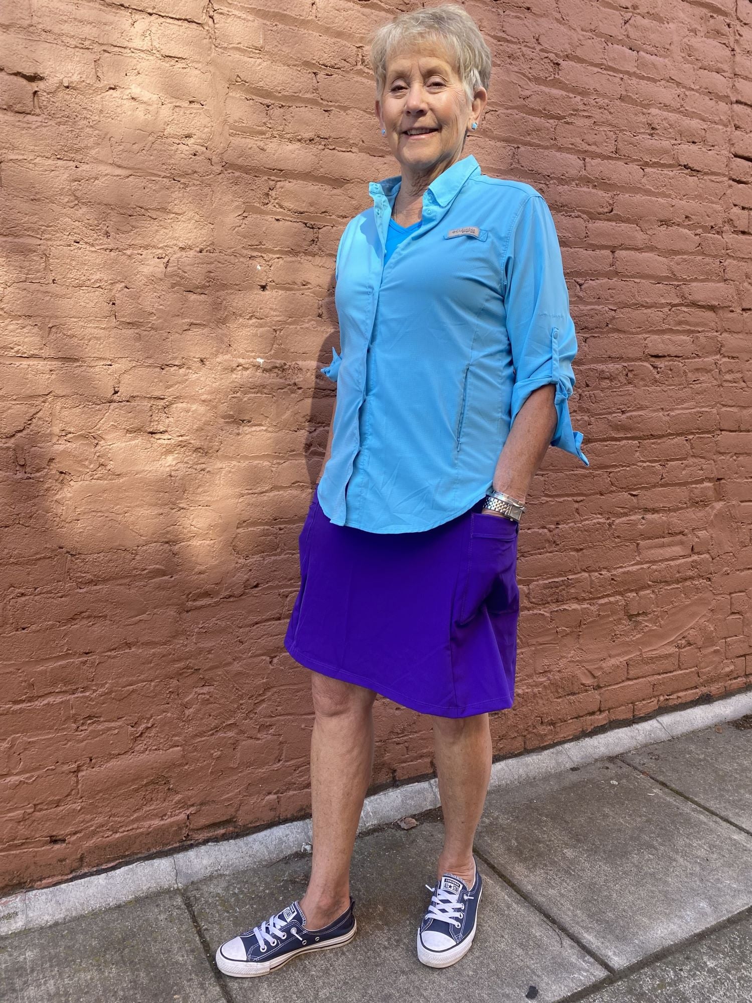 Person wearing a blue shirt and purple skirt standing against a brick wall.