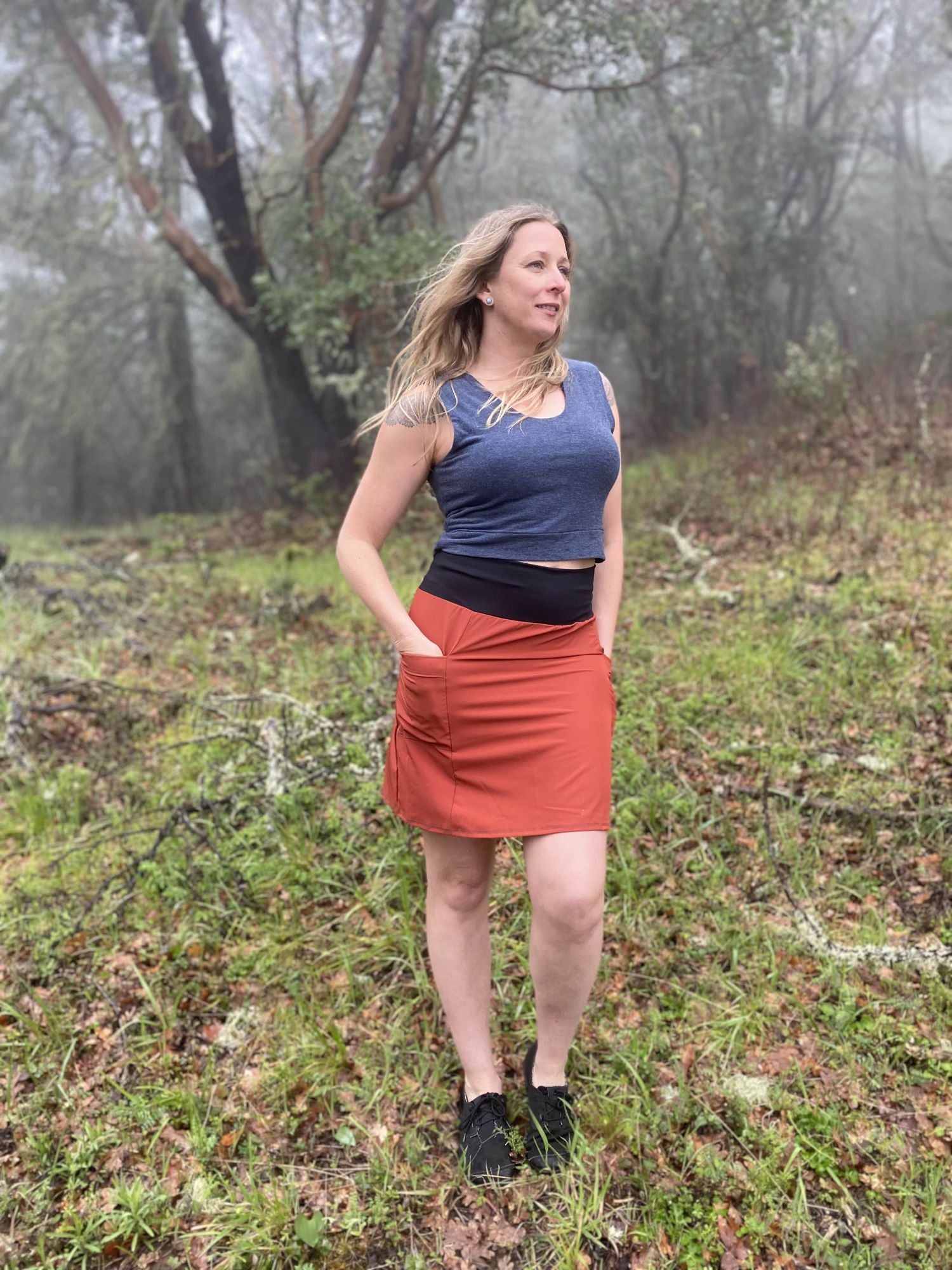 Woman standing in a forested area wearing a blue top and rust skirt.
