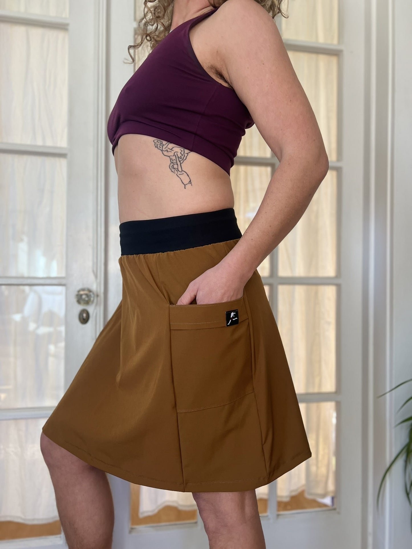 Person wearing a brown skirt kilt with a visible brand logo, standing in front of a glass door.