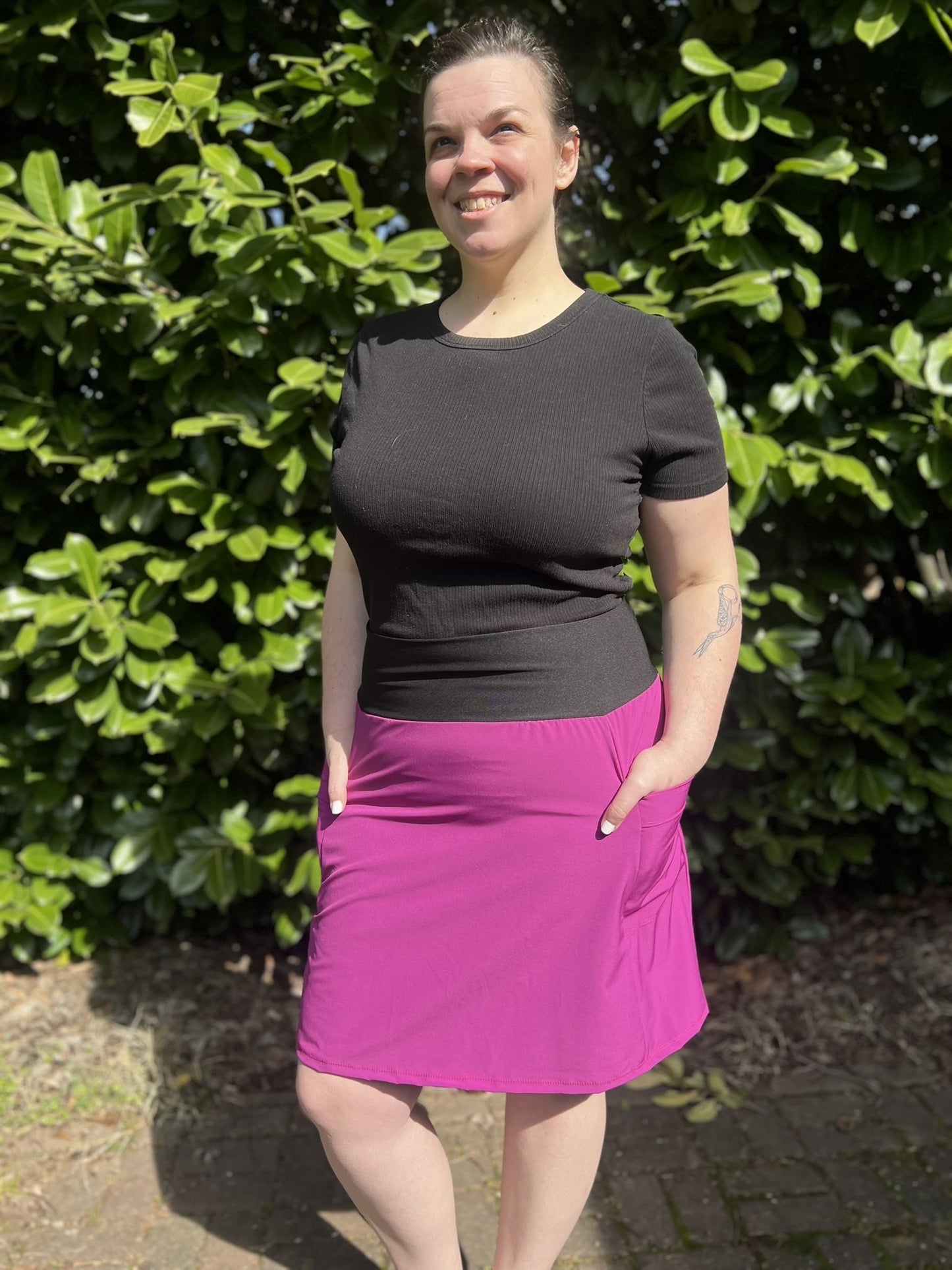 Person wearing a black top and magenta skirt standing in front of green foliage.