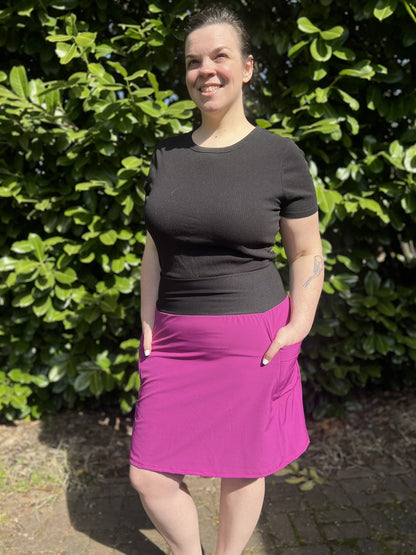 Person wearing a black top and magenta skirt standing in front of green foliage.