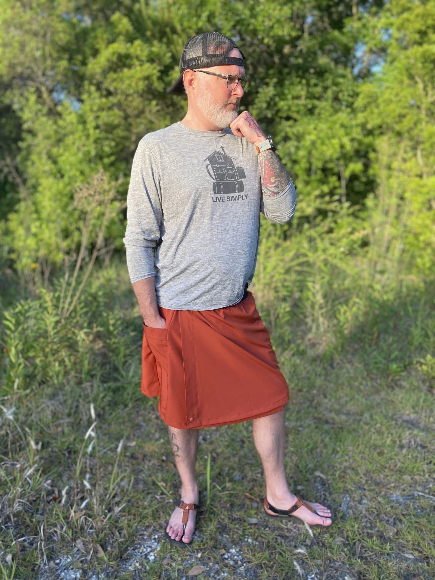 Man standing outdoors in a natural setting wearing a gray long-sleeve shirt and rust skirt
