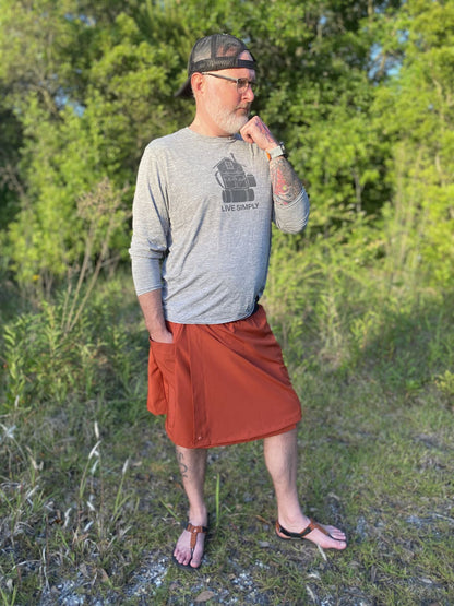 Man standing outdoors in a natural setting wearing a gray long-sleeve shirt and rust skirt
