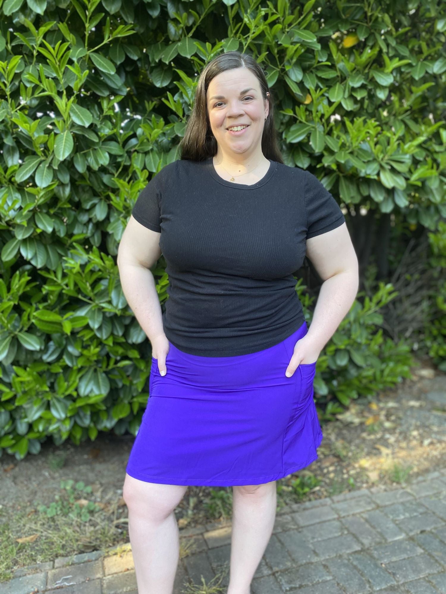 Woman wearing a black top and purple skirt standing in front of green bushes.