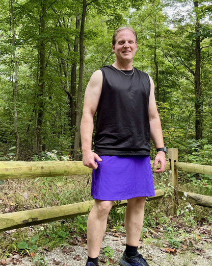 Man wearing a black sleeveless shirt and purple skirt standing in a forest.