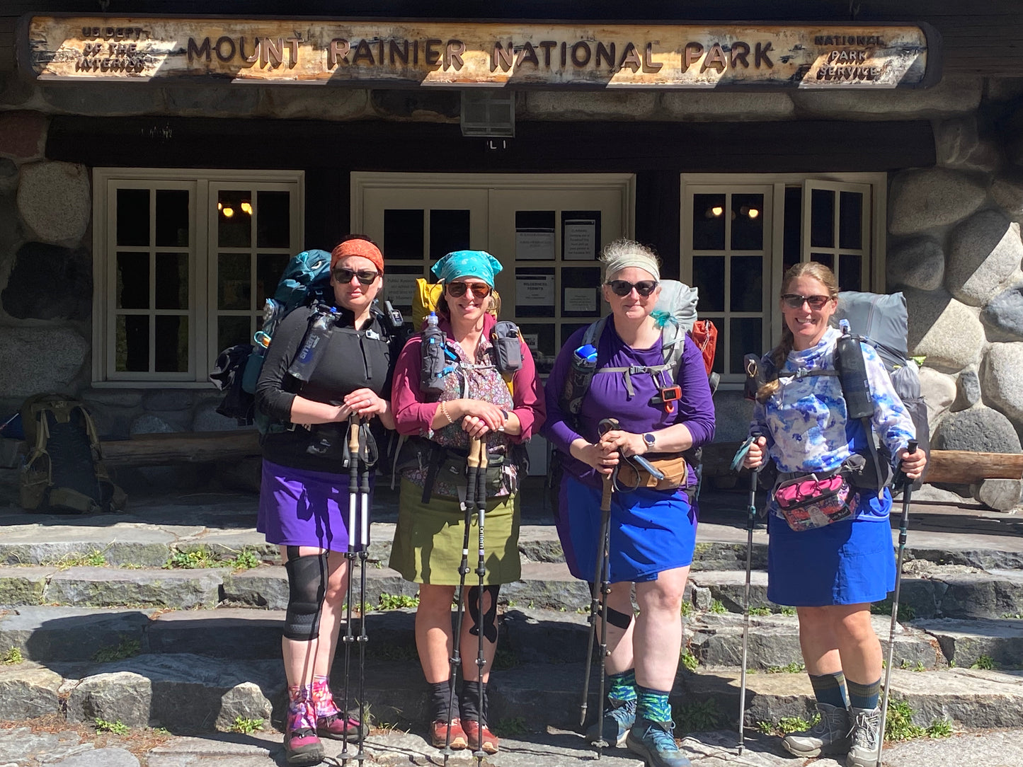 Four hikers with backpacks and sunglasses standing in front of Mount Rainier National Park entrance.