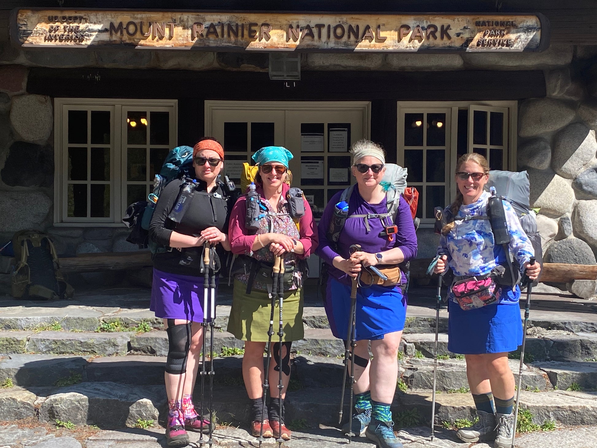 Four hikers with backpacks and sunglasses standing in front of Mount Rainier National Park entrance.